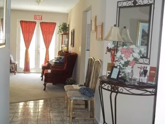 Interior view of a hallway leading to a living area with two red armchairs and a glass cabinet. The hallway has a tiled floor and two wooden chairs with cushions. On the right side, there is a decorative table with a lamp, a vase of pink and white flowers, and a large mirror above it. At the end of the hallway, there are double doors with red curtains and an exit sign above them.