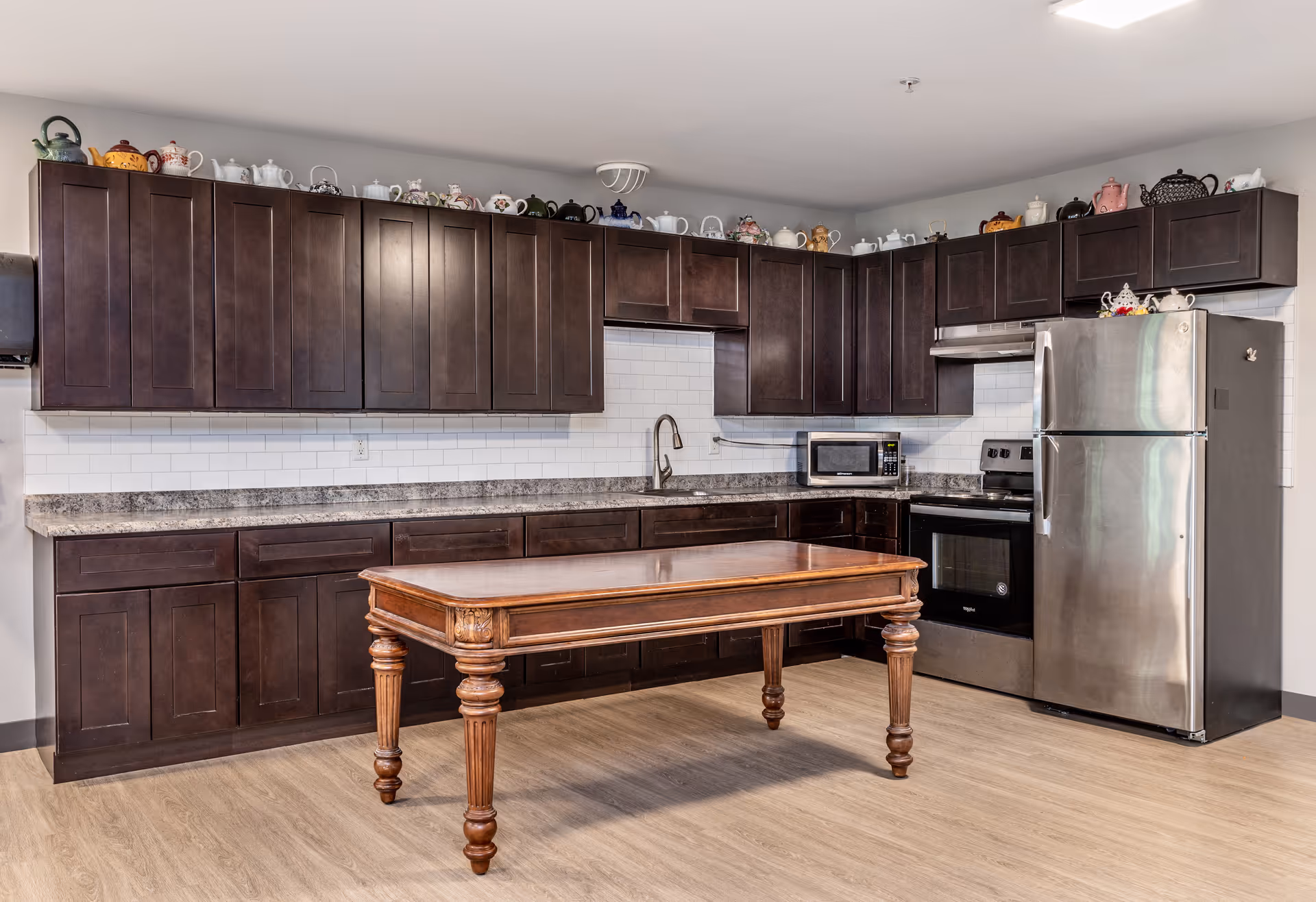 A kitchen with dark wooden cabinets, a stainless steel refrigerator and oven, a microwave on the countertop, and a wooden table with ornate legs in the center. The backsplash is white subway tile, and there is a collection of decorative teapots displayed on top of the upper cabinets.