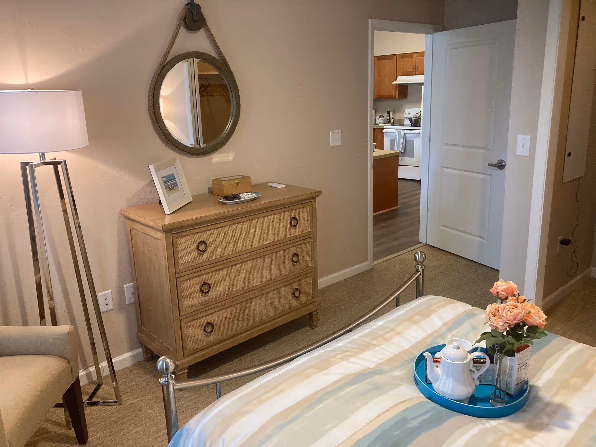 A cozy bedroom with a metal bed frame and a striped bedspread. On the bed is a blue tray holding a white teapot, cups, and a vase with pink roses. To the left is a wooden dresser with three drawers, a framed photo, a small box, and a remote control on top. Above the dresser hangs a round mirror with a rope frame. A tall floor lamp and a beige armchair are positioned to the left. The bedroom door is open, revealing a kitchen with wooden cabinets and white appliances.