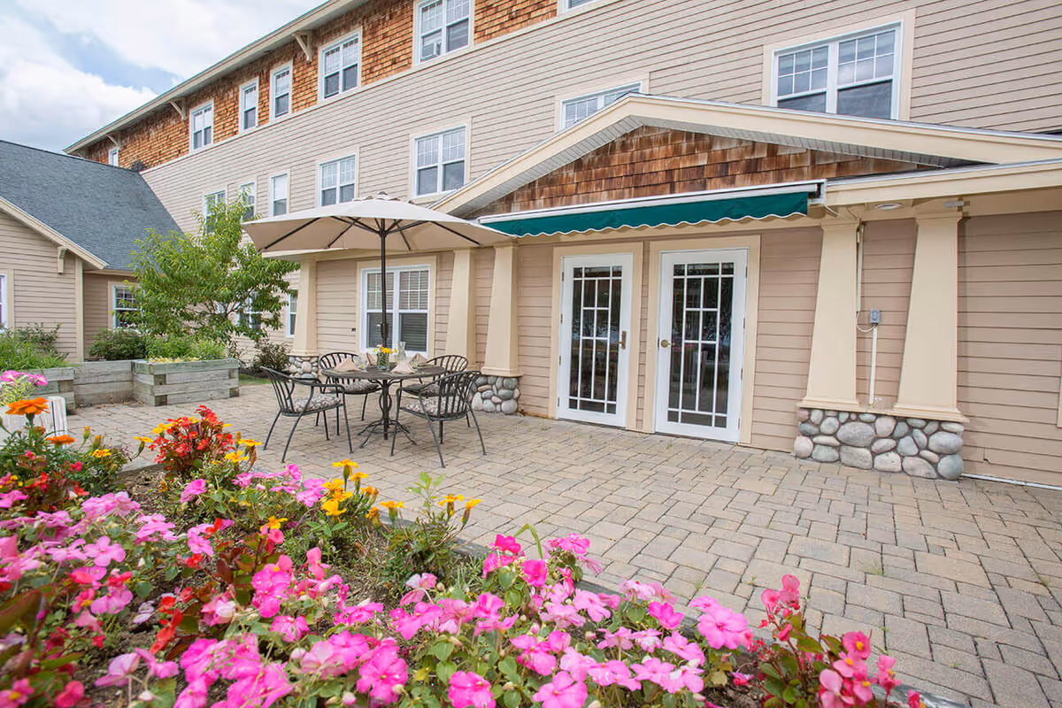 Patio courtyard with a table, umbrella and chairs surrounded by flower beds in front of a multi-story senior living building.