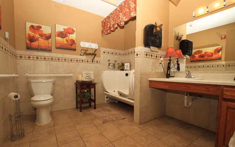 A spacious bathroom with beige tiled walls and floor, featuring a white toilet, a walk-in bathtub with a door, a wooden side table with decorative items, a large mirror above a wooden vanity with a sink, and a red floral valance above the window. The walls are decorated with floral artwork and a sign that says 'Family'.