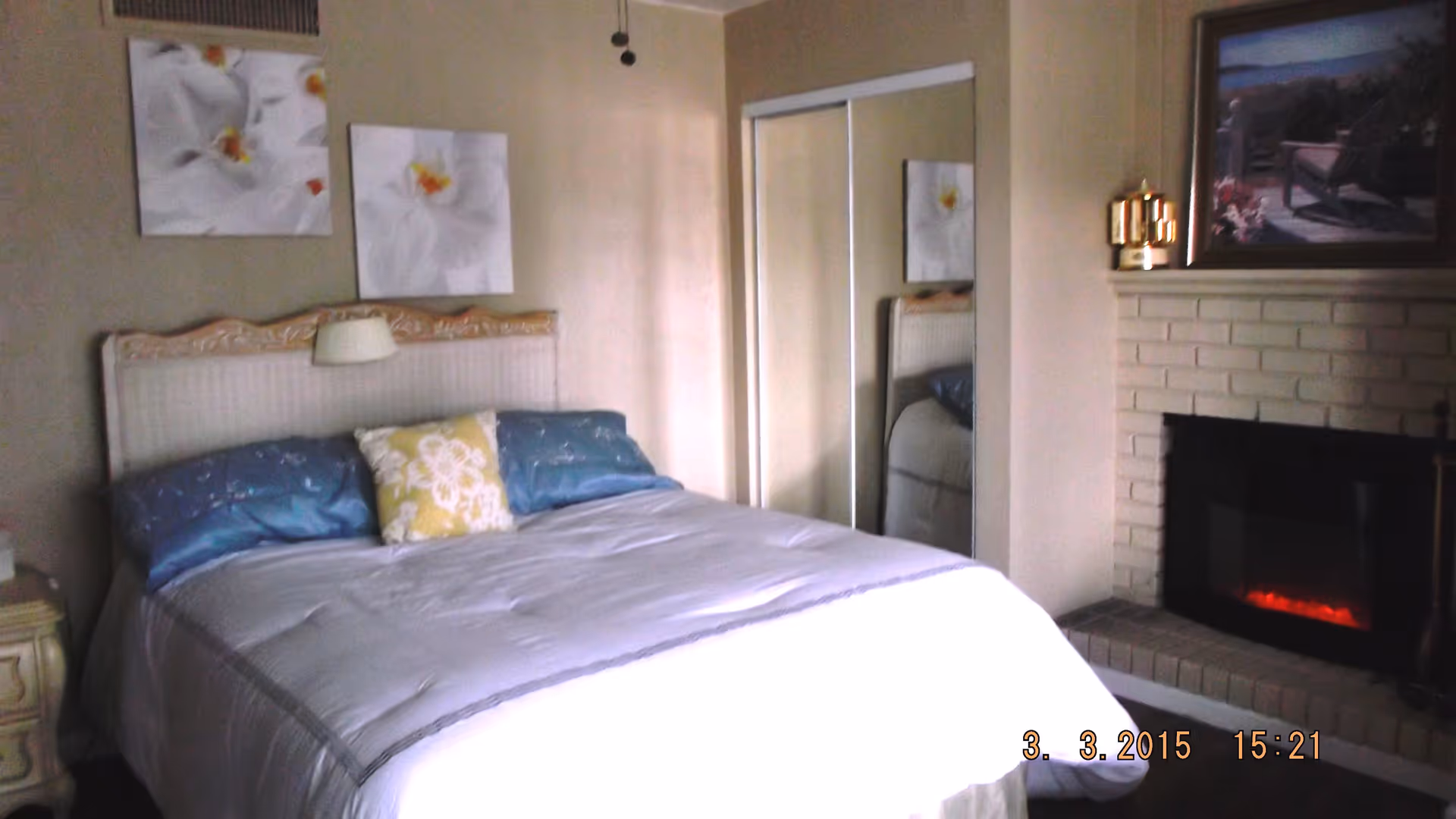 A bedroom with a large bed covered in a white comforter and blue and yellow pillows. There are two floral paintings on the wall above the headboard. To the right, there is a white brick fireplace with a painting and a decorative item on the mantel. A mirrored closet door is visible next to the bed.