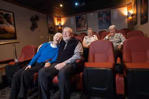 Four elderly people sitting in red theater seats inside a small movie theater room, smiling and enjoying themselves. Movie posters are visible on the walls behind them.