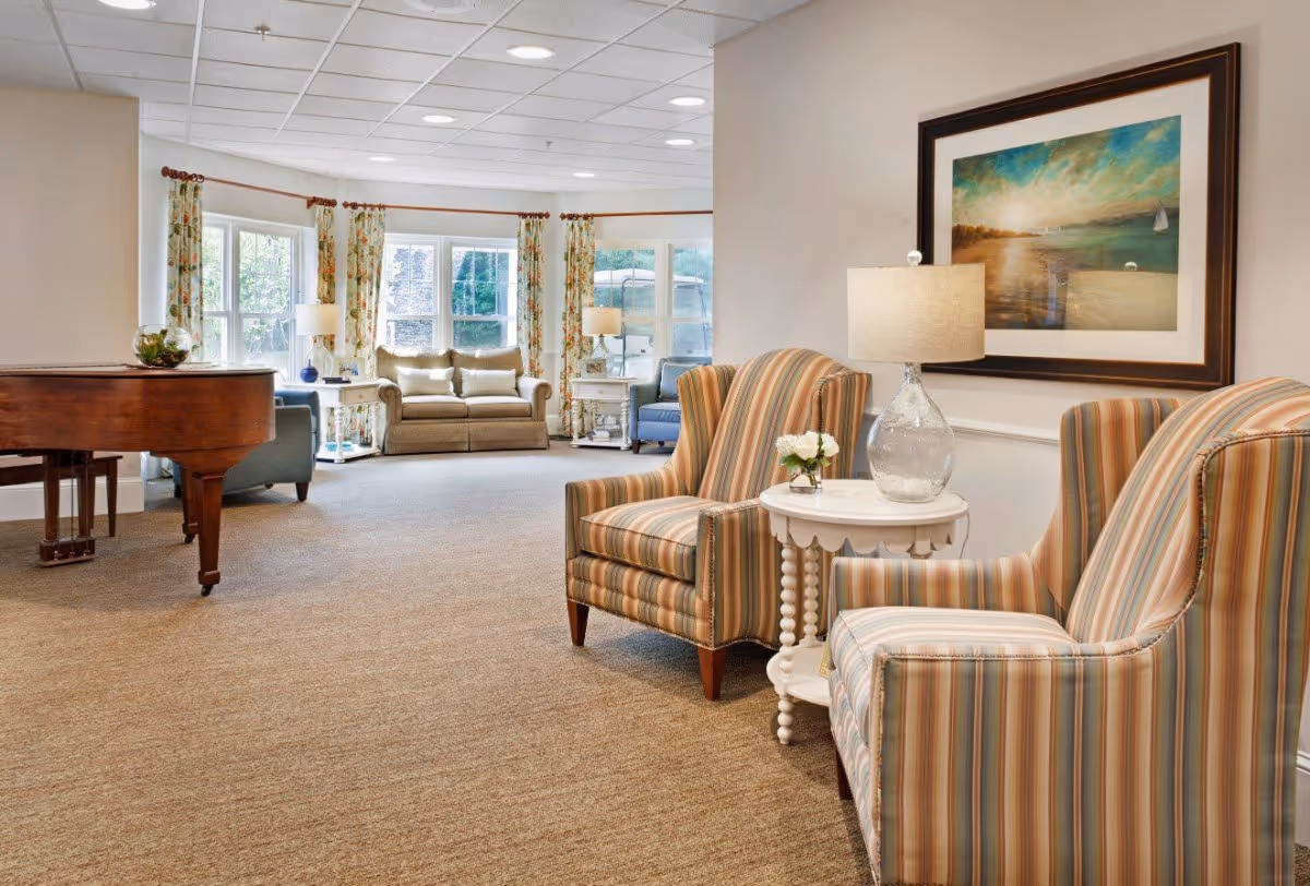Bright communal lounge with striped armchairs and a small side table, a grand piano, and a sofa by bay windows.