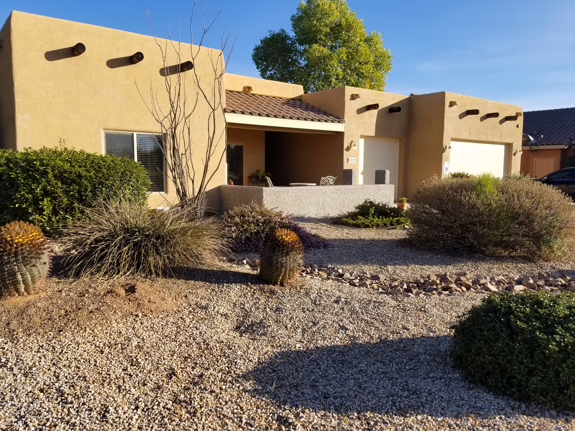 Single-story southwestern-style stucco building with gravel desert landscaping, cacti, a small covered patio and two garage doors under a clear sky.