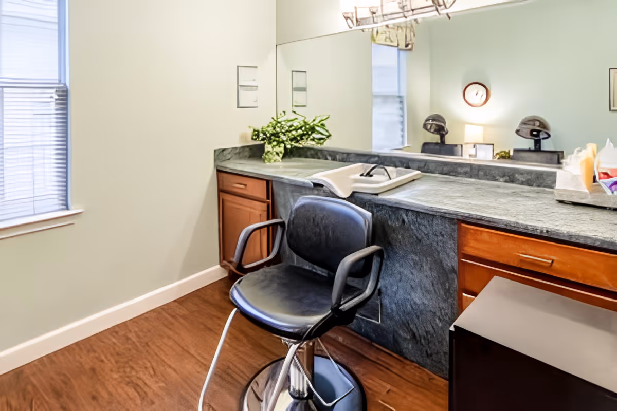 Interior view of a salon area with a black salon chair in front of a long countertop with a large mirror. The countertop has a towel, some hair care items, and a small plant. There are two hair dryer chairs visible in the mirror reflection, along with a clock on the wall and a window with blinds.