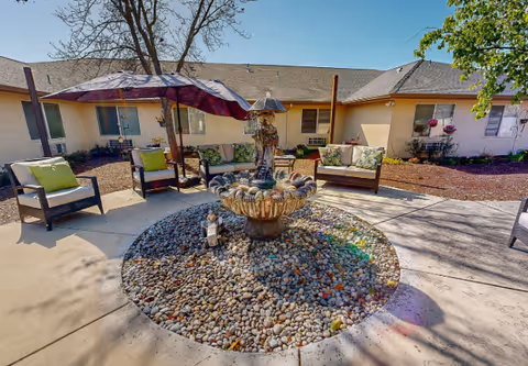 Outdoor seating area with cushioned chairs and a sofa arranged around a central decorative fountain filled with pebbles. A large umbrella provides shade, and the area is surrounded by a single-story building with windows and some plants.