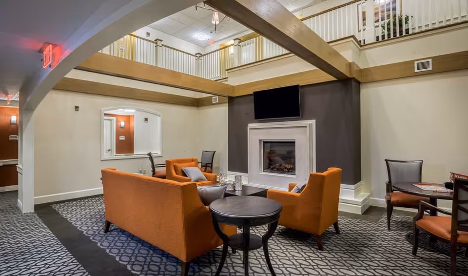 A cozy living room area in a senior living facility featuring orange upholstered chairs and a sofa arranged around a dark wood coffee table. The room has a modern fireplace with a mounted flat-screen TV above it. The space has a high ceiling with wooden beams and a second-floor balcony railing visible. The carpet has a geometric pattern, and there is a small round table with chairs in the corner.