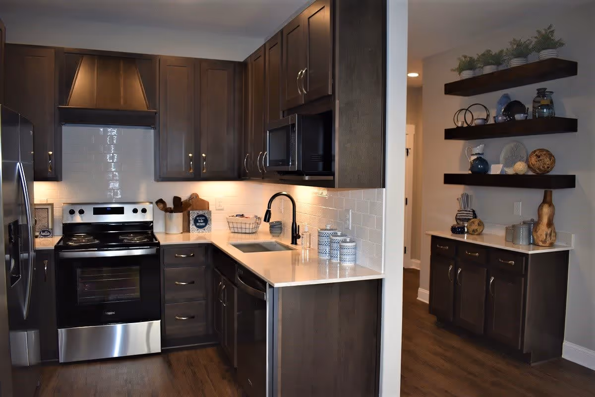 Modern kitchen with dark wood cabinets, stainless steel appliances including a refrigerator, oven, and microwave. The kitchen features a white countertop with a black faucet and sink, white subway tile backsplash, and decorative shelves with plants and pottery on the adjacent wall.