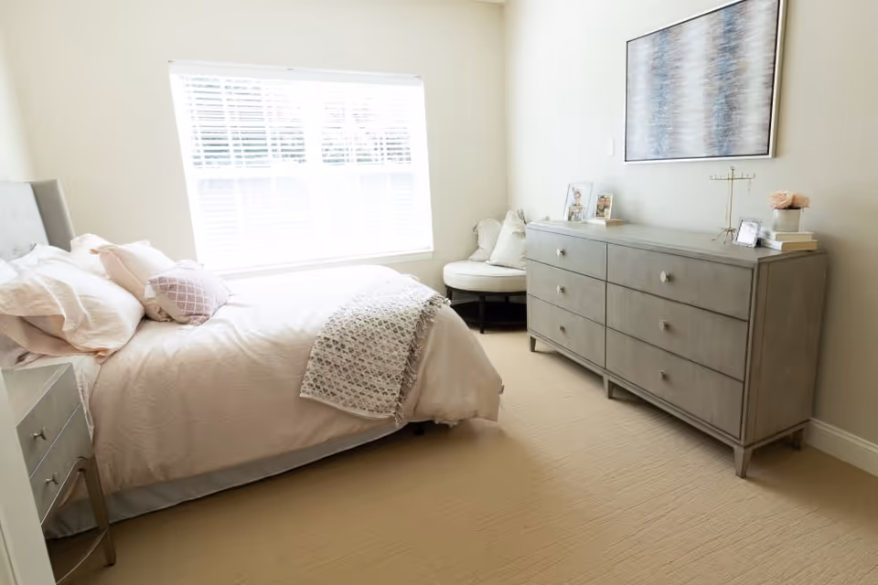 Sunlit bedroom with a neatly made bed, a six-drawer dresser, and a round accent chair by a large window.