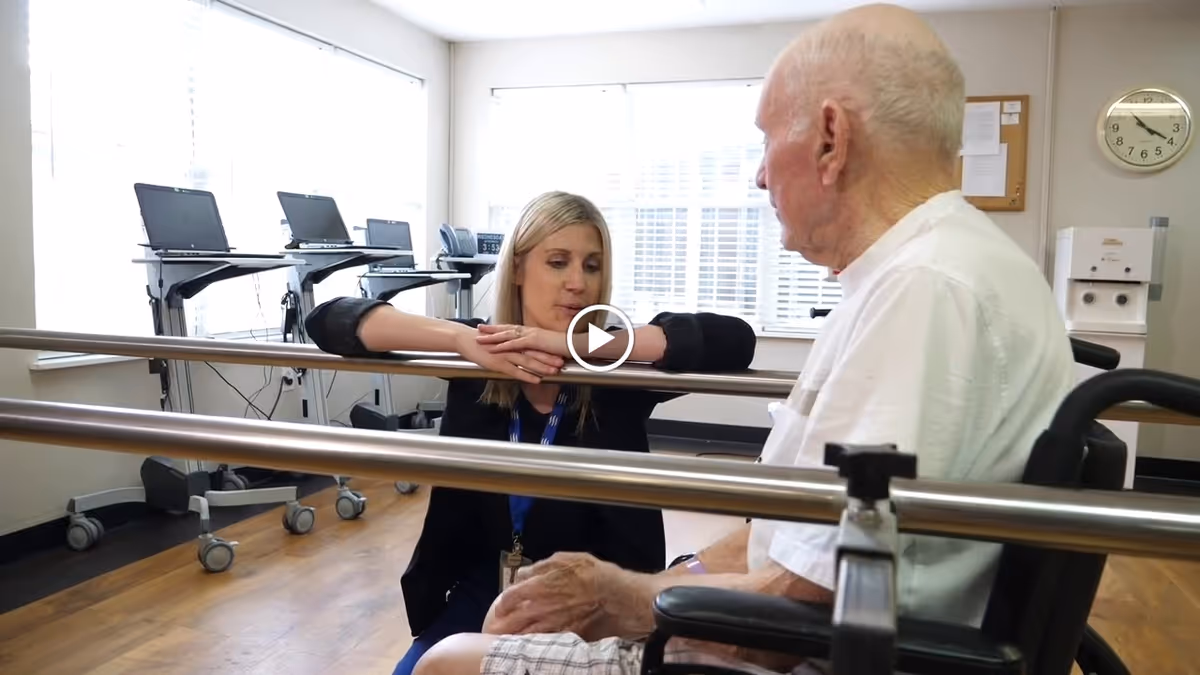 A healthcare professional is kneeling and talking to an elderly man seated in a wheelchair in a rehabilitation room with parallel bars and exercise equipment in the background.
