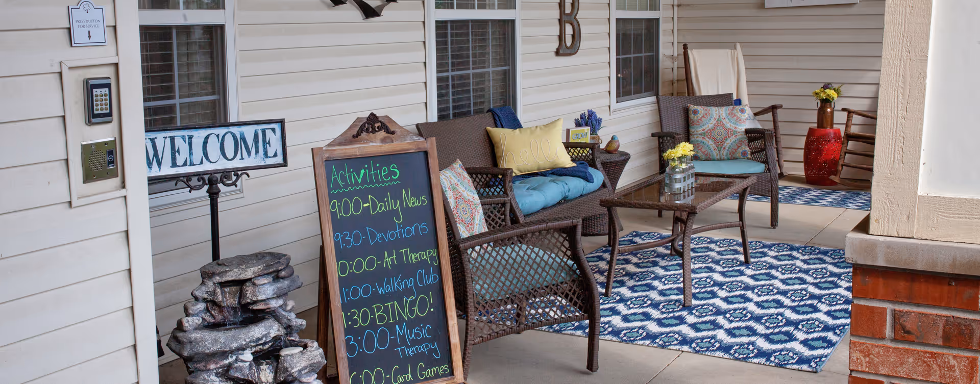 Outdoor seating area at a senior living community with wicker chairs and a loveseat adorned with colorful cushions and pillows. A glass-top coffee table sits on a blue patterned rug. A chalkboard sign lists daily activities including Daily News, Devotions, Art Therapy, Walking Club, Bingo, Music Therapy, and Card Games. A welcome sign and a small water fountain are also visible near the entrance door.