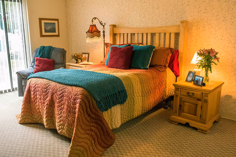 A cozy bedroom with a wooden bed frame, a multicolored quilt, and several pillows in red and teal. There is a wooden nightstand with a lamp, flowers, and framed photos. A comfortable armchair with a red pillow and teal throw is placed near a window with vertical blinds. The walls have a light floral wallpaper and a framed picture.