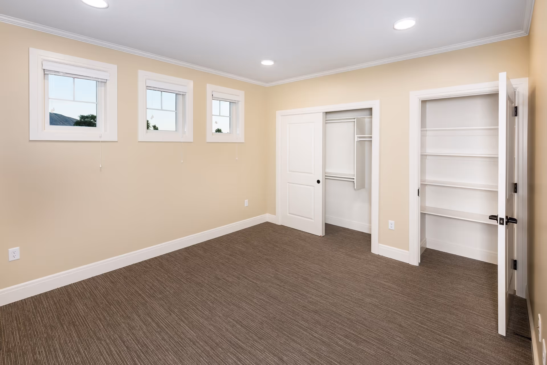 Empty room with beige walls, three small windows with white frames, brown carpeted floor, a closet with sliding white doors, and an open door revealing white built-in shelves.