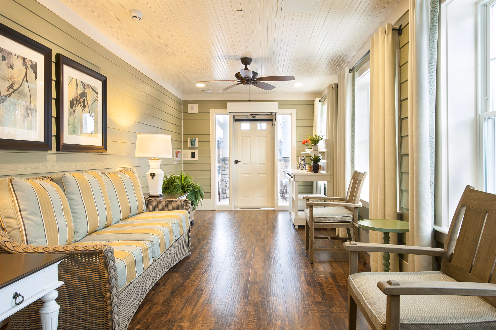 A bright and cozy sitting area with a striped cushioned wicker sofa on the left, two wooden armchairs with cushions on the right, a small green side table, and a white door at the end of the room. The walls are painted light green with horizontal paneling, and there are two framed artworks above the sofa. The floor is wooden, and the ceiling has a fan with lights. Large windows with beige curtains let in natural light.