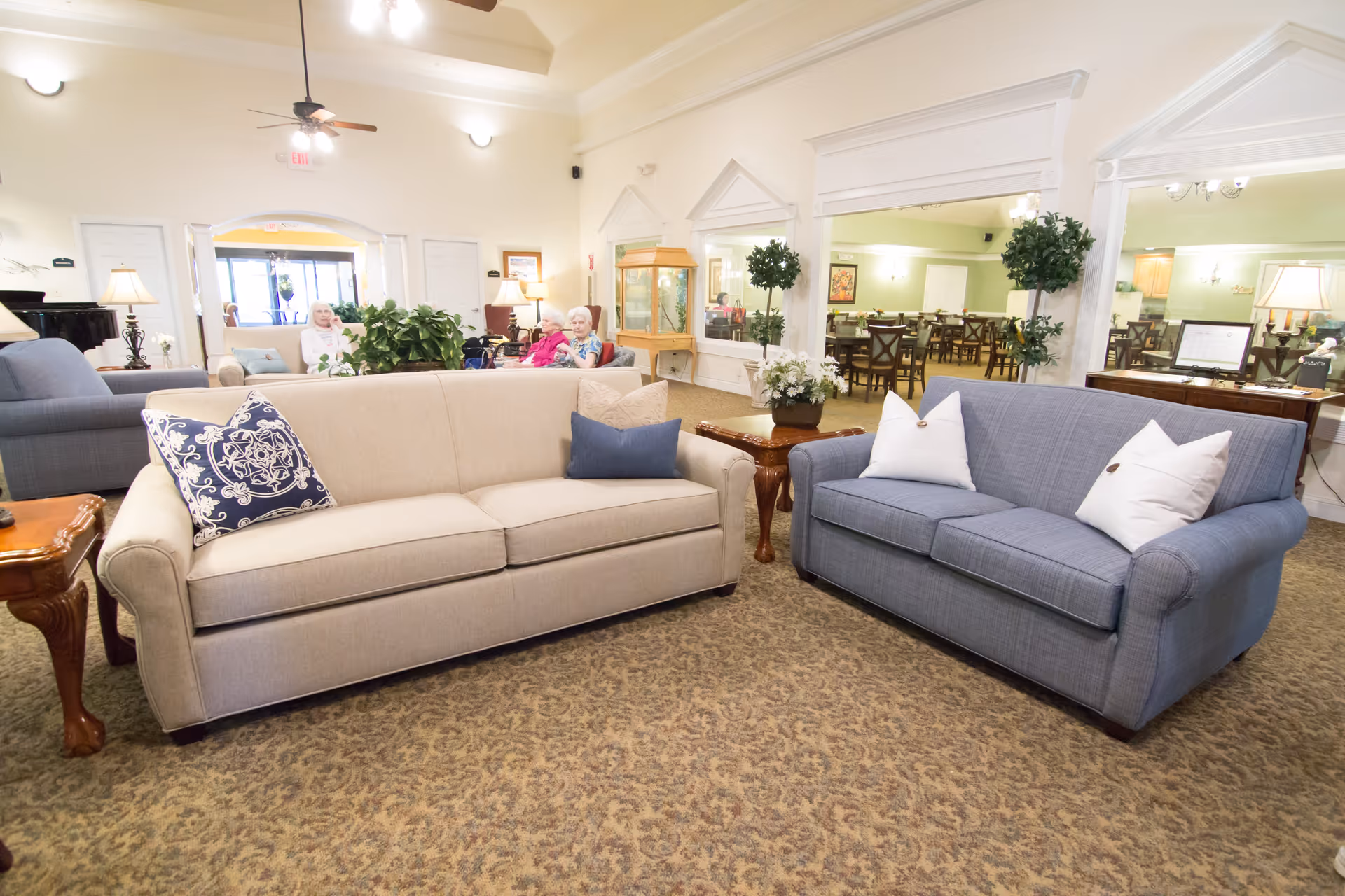 A bright and spacious living room area in a senior living facility with two sofas, one beige and one blue, each adorned with decorative pillows. There are wooden side tables with lamps and plants, and in the background, a dining area with tables and chairs is visible through large interior windows. Two elderly women are seated in the background near the entrance.