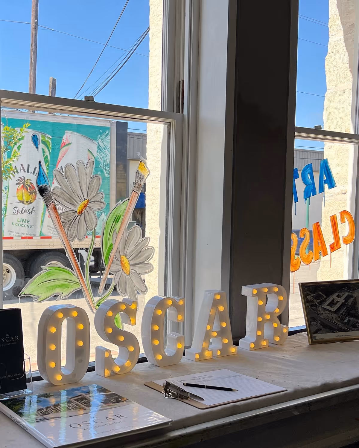 A window sill display featuring illuminated letters spelling 'OSCAR' with small light bulbs inside each letter. Behind the letters, there are painted flowers and paintbrushes on the window glass. On the sill, there is a clipboard with a pen, a framed photo, and a brochure or booklet titled 'OSCAR'. Outside the window, a truck with a colorful advertisement is visible.