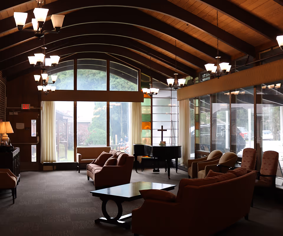 A spacious living room with high wooden beam ceilings and multiple chandeliers. The room features several upholstered sofas and armchairs arranged around a central coffee table. Large windows and glass walls allow natural light to fill the space, with a view of greenery and parked cars outside. A black grand piano with a cross on top is positioned near the windows.