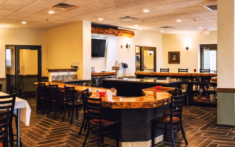 Interior view of a dining area in The Meadows Of Fond Du Lac featuring a U-shaped wooden bar with several black chairs around it. The room has warm lighting, a tiled floor with a herringbone pattern, and tables with chairs covered in white tablecloths. There is a TV mounted on the wall and various kitchen and dining supplies visible in the background.