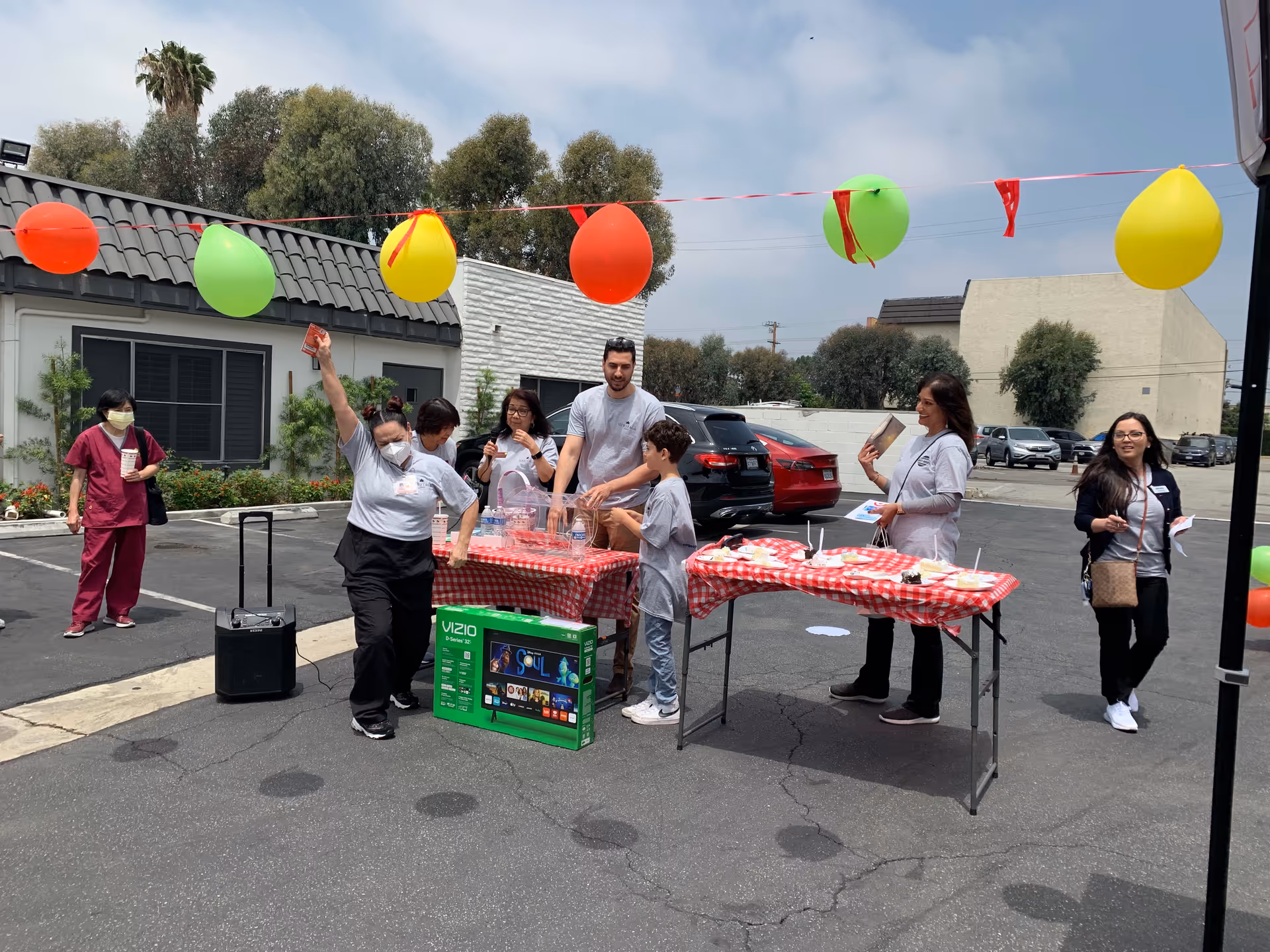 A group of people gathered outdoors in a parking lot area decorated with colorful balloons. They are standing around two tables covered with red and white checkered tablecloths, with snacks and drinks on the tables. One person is holding up a card, and there is a boxed Vizio television on the ground near the tables. Trees and buildings are visible in the background under a partly cloudy sky.