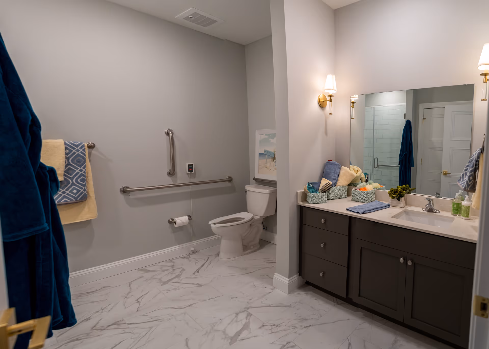 Bright accessible bathroom with toilet and grab bars, marble-look floor, and a vanity with sink and mirror.