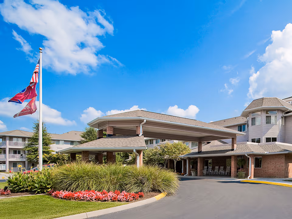 Exterior view of Holiday Echo Ridge senior living facility showing the main entrance with a covered drop-off area, landscaped flower beds, and flags on a flagpole under a bright blue sky with some clouds.
