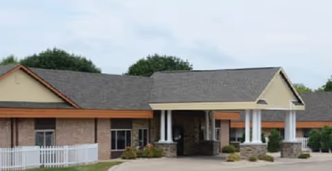 Exterior view of a single-story senior living facility building with a covered entrance supported by white columns, beige walls, and a gray shingled roof. There is a white fence on the left side and some greenery around the building.
