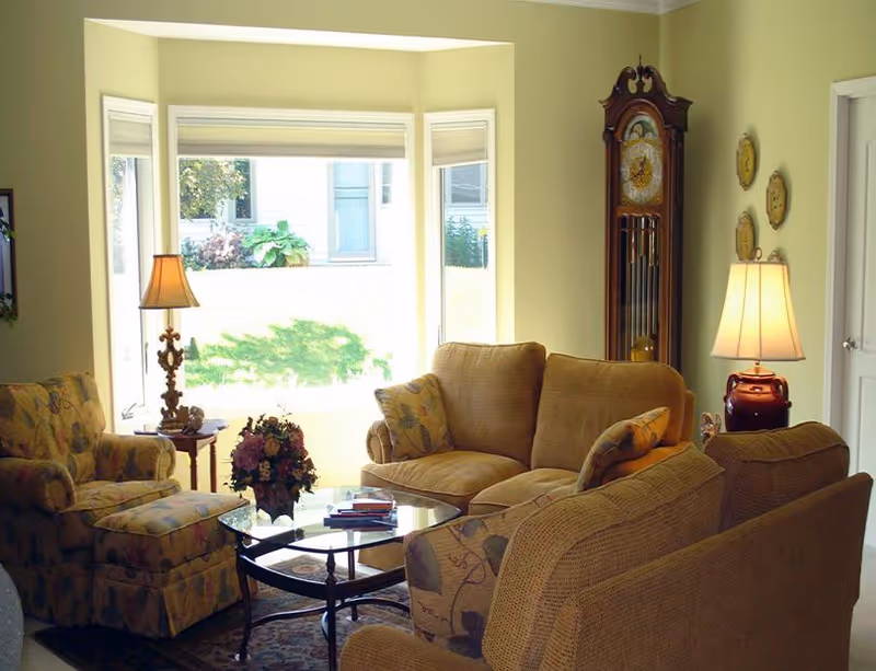 A cozy living room with beige and floral upholstered sofas and armchairs arranged around a glass coffee table with a flower arrangement. There are two table lamps on either side, a tall grandfather clock in the corner, and a large window letting in natural light.