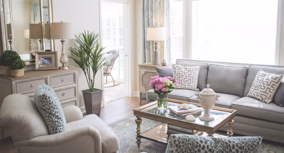 A bright and cozy living room with a gray sofa adorned with patterned pillows, a glass coffee table with a vase of pink flowers and books, a beige armchair with a patterned cushion, a wooden dresser with a lamp, framed photo, and a potted plant, and a tall green plant in a square pot near a doorway leading to a sunlit room with wicker furniture.