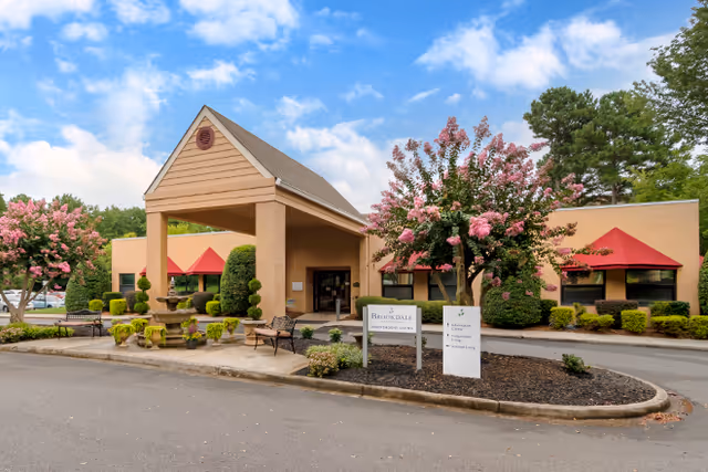 Exterior view of Brookdale Charlotte East senior living facility with a beige building featuring a covered entrance, red awnings over windows, landscaped bushes and flowering trees, and a sign displaying the facility name and services offered.