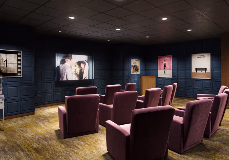 A small movie theater room with dark paneled walls and a carpeted floor. There are two rows of plush purple armchairs facing a screen showing a black and white film scene. Movie posters are displayed on the walls, and a wooden podium stands near the back.