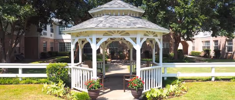 A white wooden gazebo with a shingled roof situated in a green courtyard area with trees and a white fence. The background shows a multi-story brick building with windows and archways.