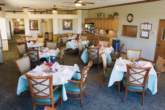 A dining room with multiple tables covered in light blue tablecloths and white napkins, each set with cups and floral centerpieces. Wooden chairs with cushioned seats surround the tables. The room has ceiling fans, framed artwork on the walls, and a wooden cabinet area with a countertop and various items on top.