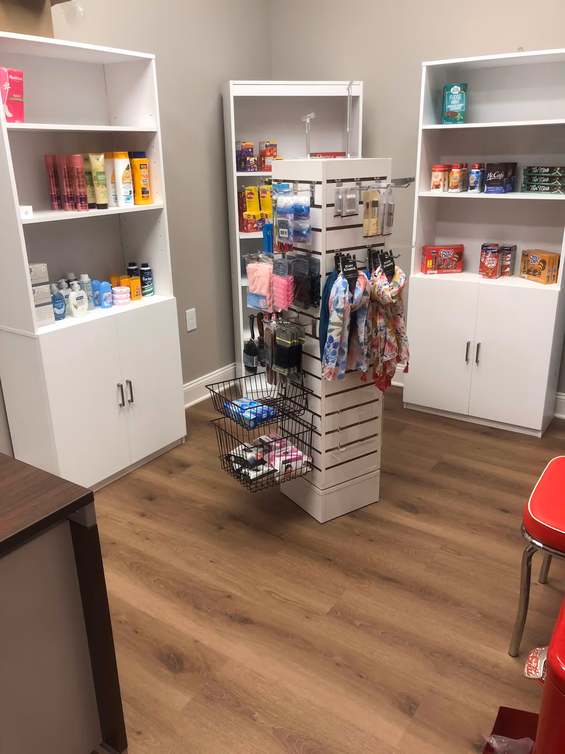 Interior view of a small retail or convenience area with white shelves and a central display rack. The shelves contain personal care products, snacks, and other small items. The floor is wood, and a red chair is partially visible on the right side.