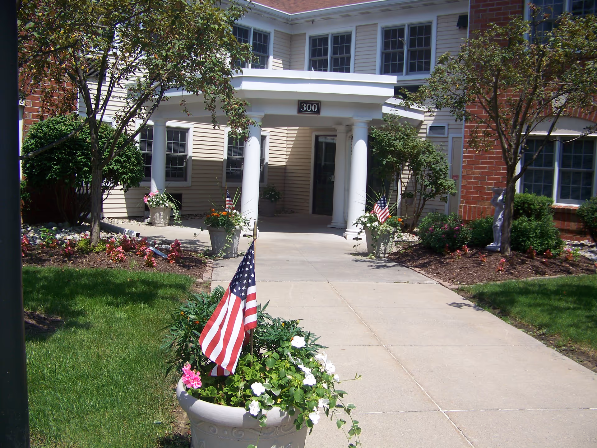 Entrance to a building with white columns and a covered porch, surrounded by landscaped gardens with flowers and small American flags in planters. The building has beige siding and red brick walls with multiple windows.