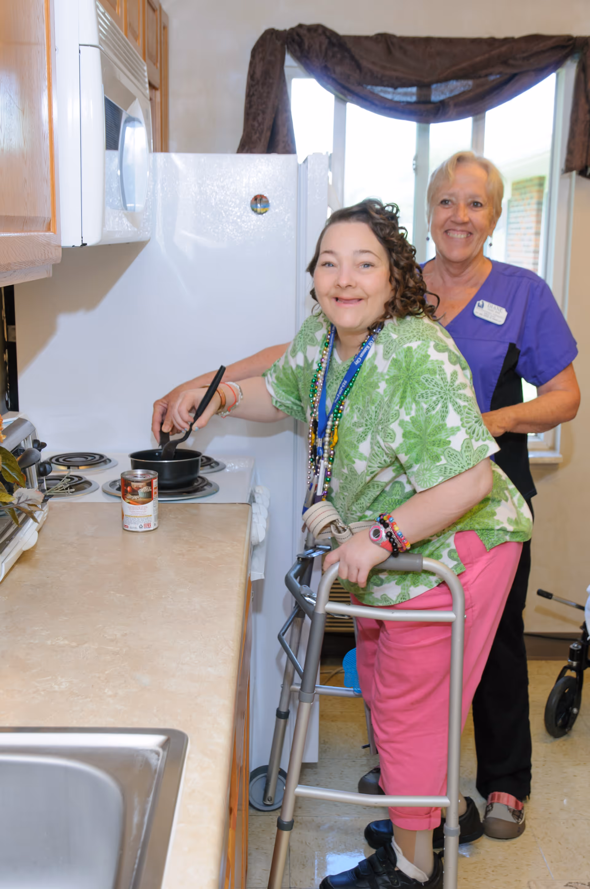 A woman using a walker is cooking on a stove in a kitchen, assisted by a smiling caregiver standing behind her. The kitchen has wooden cabinets, a white stove, and a countertop with a can of food on it.