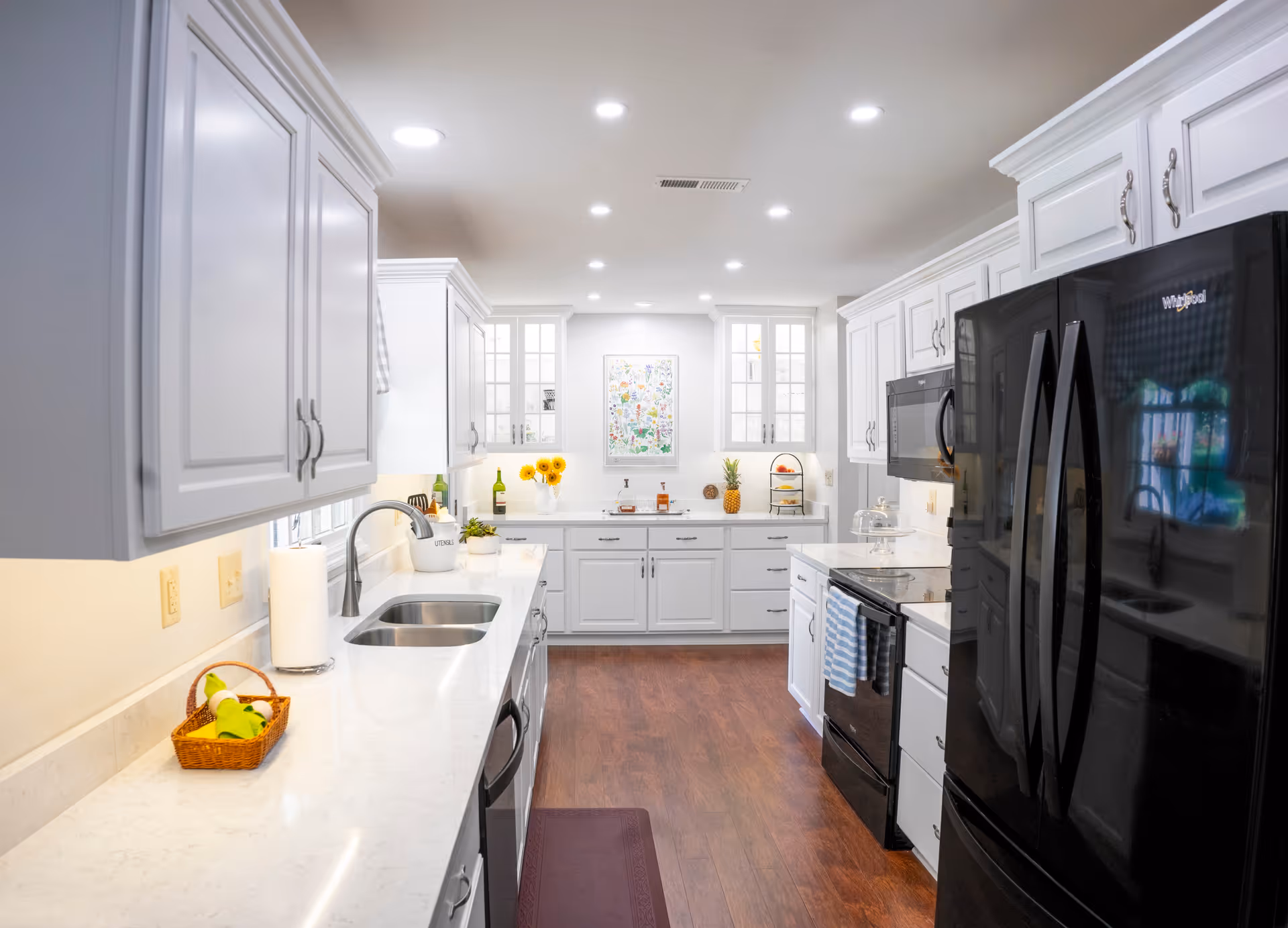 Bright and modern kitchen with white cabinets, marble countertops, a double sink, black Whirlpool refrigerator, black stove, microwave, and wooden flooring. The kitchen is well-lit with recessed ceiling lights and has decorative items including sunflowers, a pineapple, and a framed picture on the far wall.