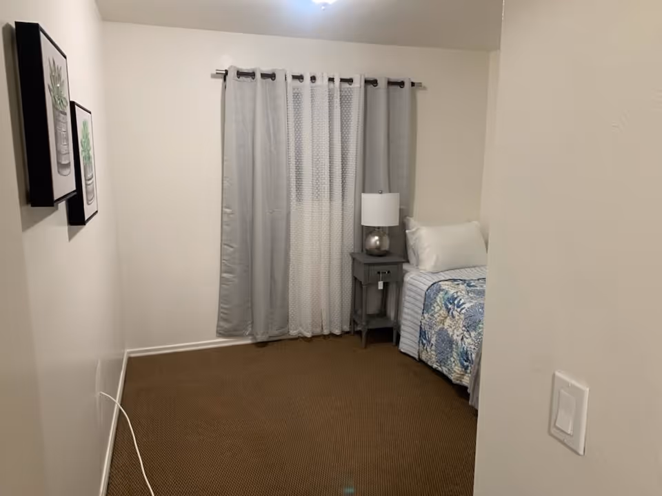 A small bedroom with a single bed covered with a blue and white floral bedspread. Next to the bed is a gray nightstand with a silver lamp on it. The window behind the bed has gray curtains with a sheer white curtain in the middle. Two framed pictures of plants hang on the left wall. The floor is carpeted in brown, and a light switch is visible on the right wall near the entrance.