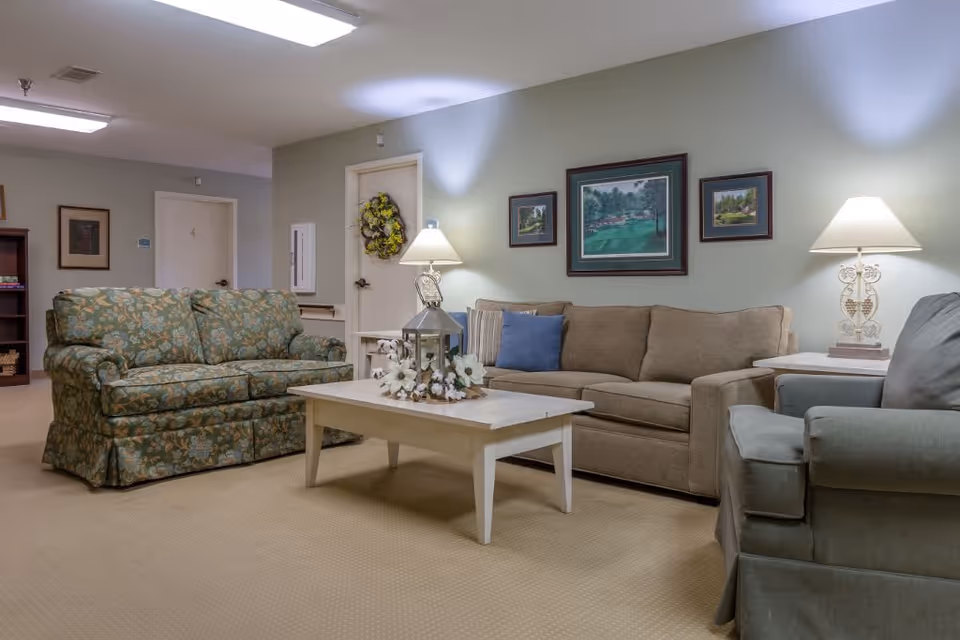 A cozy living room area in an assisted living facility featuring a floral patterned loveseat, a beige sofa with a blue pillow, and a gray armchair. A white coffee table with a decorative lantern and flowers sits in the center. Two table lamps illuminate the room, and three framed landscape paintings hang on the light green wall behind the sofa. A door with a wreath and another door labeled with the number 4 are visible in the background.