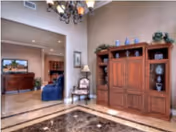 Spacious interior room with polished marble floor and a large wooden cabinet against the wall. The room features a chandelier hanging from the ceiling, a decorative chair with a lamp beside it, and an open doorway leading to another room with furniture including a blue sofa and a wooden dresser.
