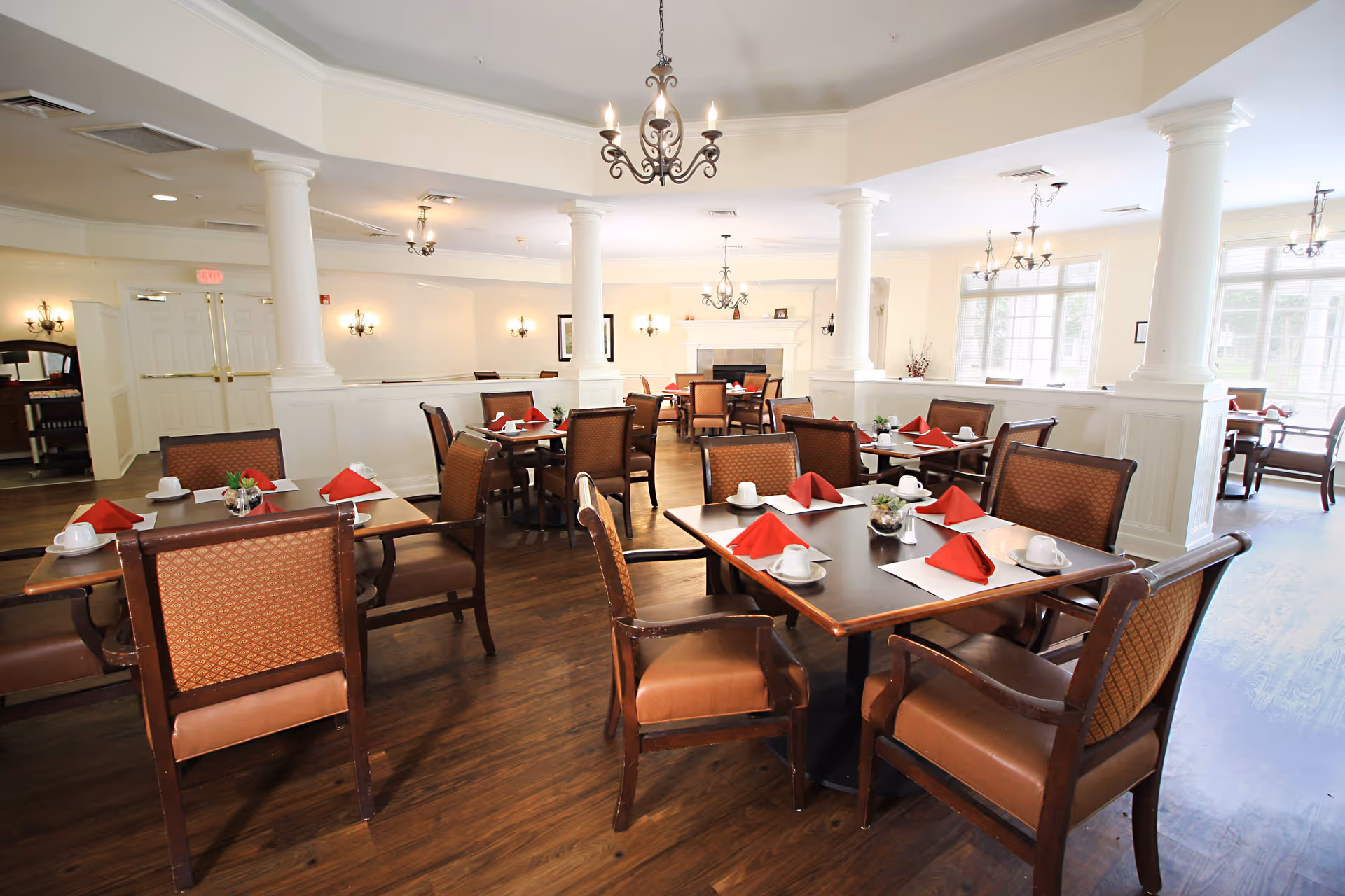 A bright and elegant dining room with multiple wooden tables and cushioned chairs. Each table is set with white cups, saucers, and red folded napkins. The room features white walls, large windows allowing natural light, decorative columns, chandeliers, and a fireplace in the background.