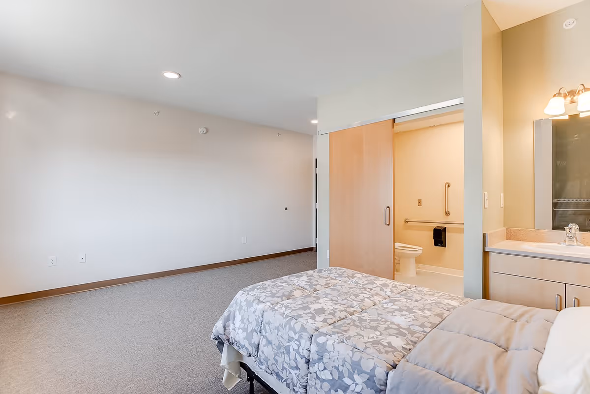 Bedroom with a bed in the foreground and an open accessible bathroom with a sliding door and vanity visible.