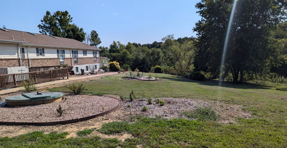Outdoor view of a senior living facility showing a grassy yard with landscaped areas, a circular stone feature, and a two-story building with brick and siding in the background under a clear blue sky.