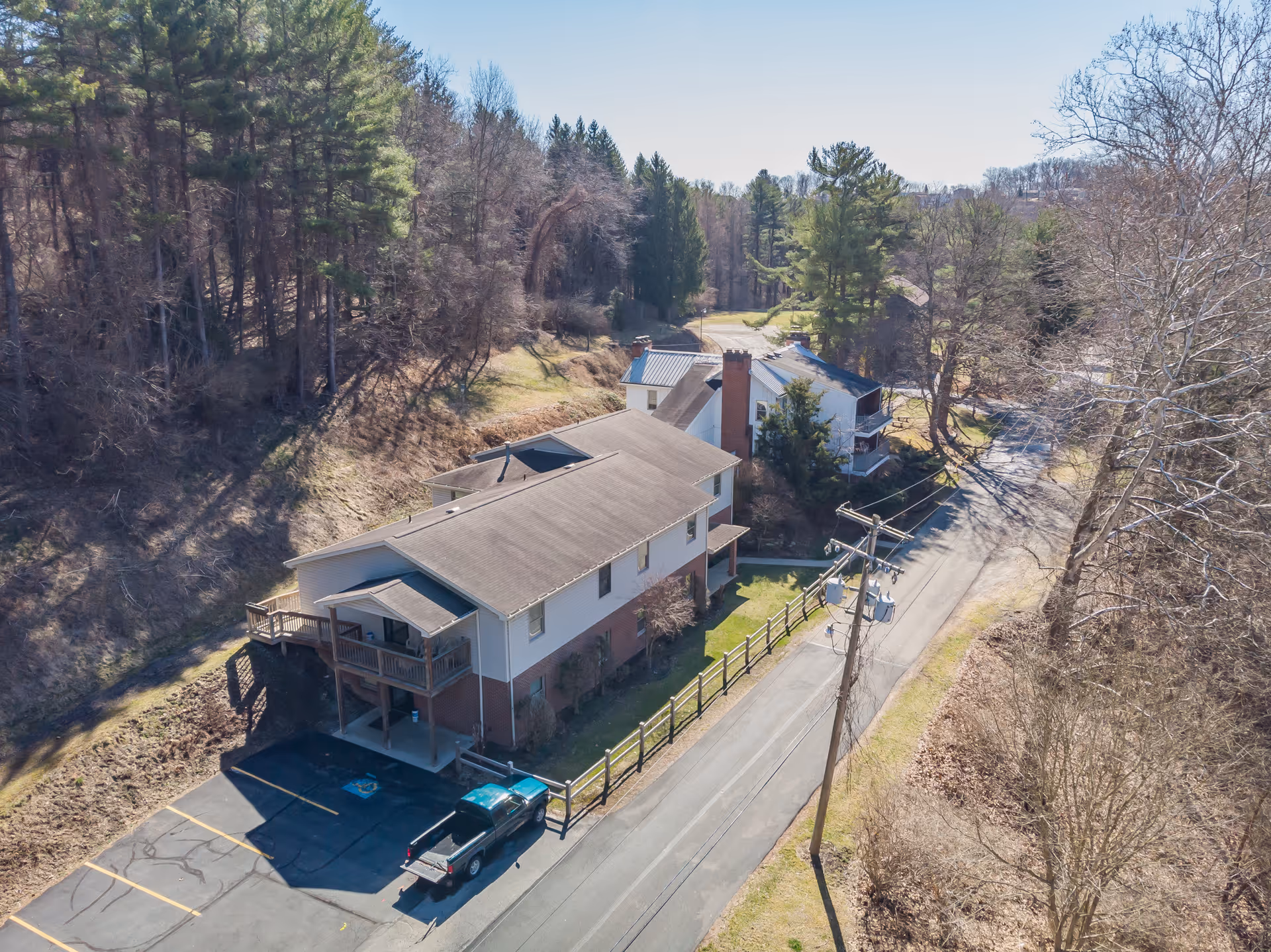 Aerial view of a two-story building with a parking lot and a pickup truck parked in front. The building is surrounded by trees and situated along a narrow road. The area appears to be in a rural or wooded setting with clear skies.