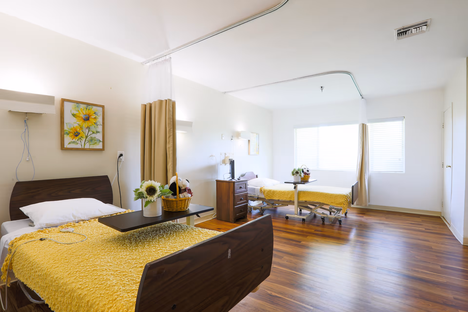 A bright and clean healthcare facility room with two hospital beds, each covered with yellow blankets. The room has wooden flooring, white walls, and large windows letting in natural light. There are bedside tables, a painting of sunflowers on the wall, and privacy curtains between the beds.