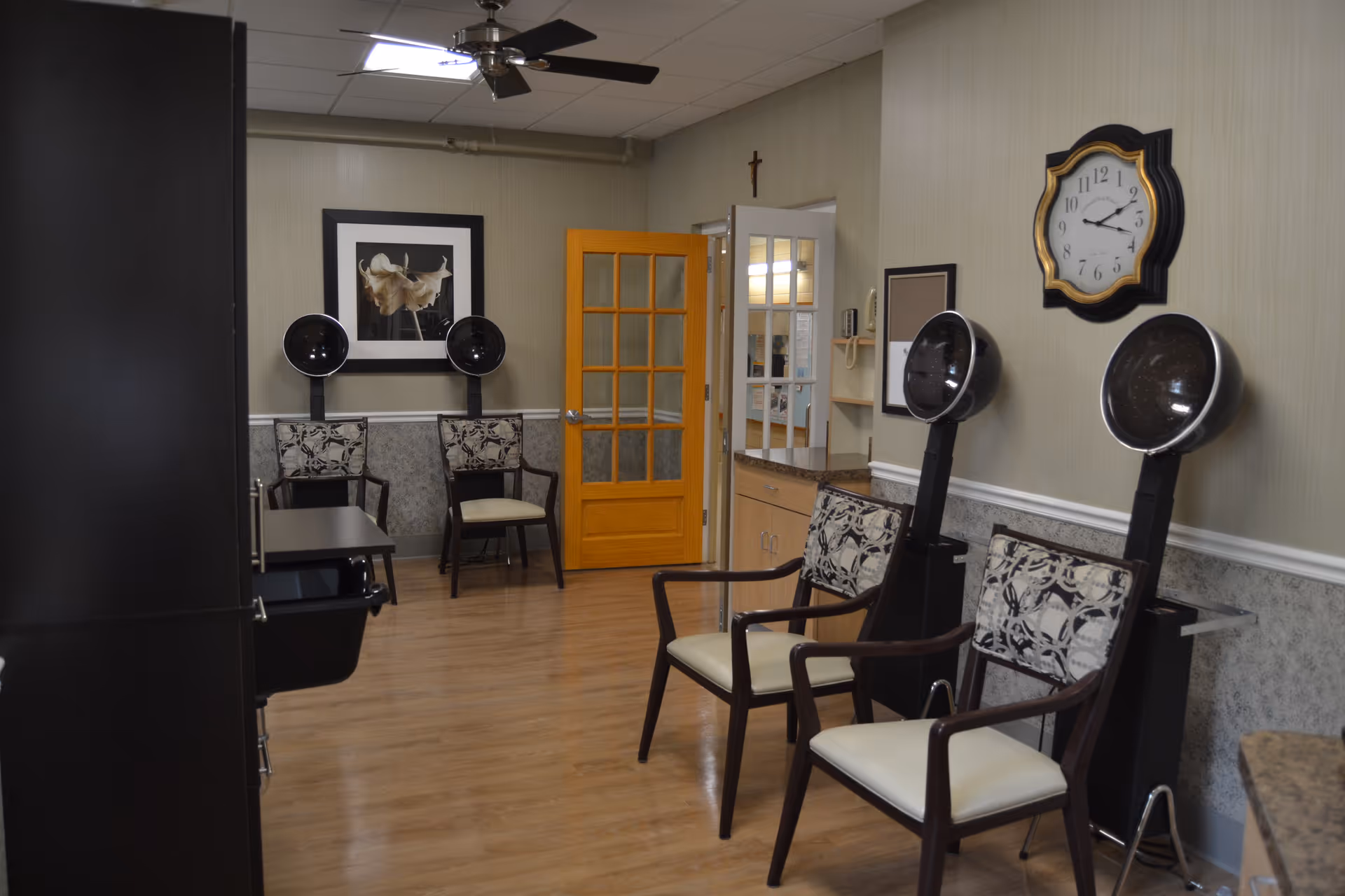 Small salon area in a nursing facility with chairs and hooded hair dryers, a wall clock, and a yellow door.