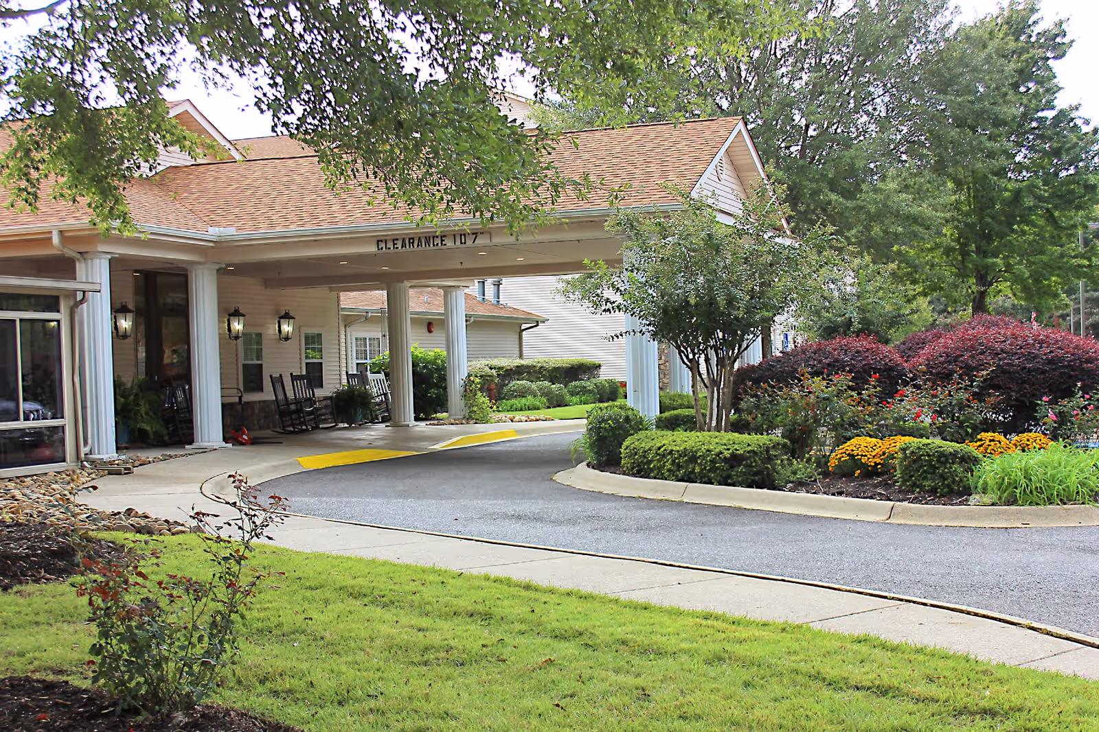 Entrance area of a senior living community building with a covered driveway supported by white columns. There are rocking chairs on the porch, well-maintained landscaping with green grass, bushes, and colorful flowers, and trees providing shade.