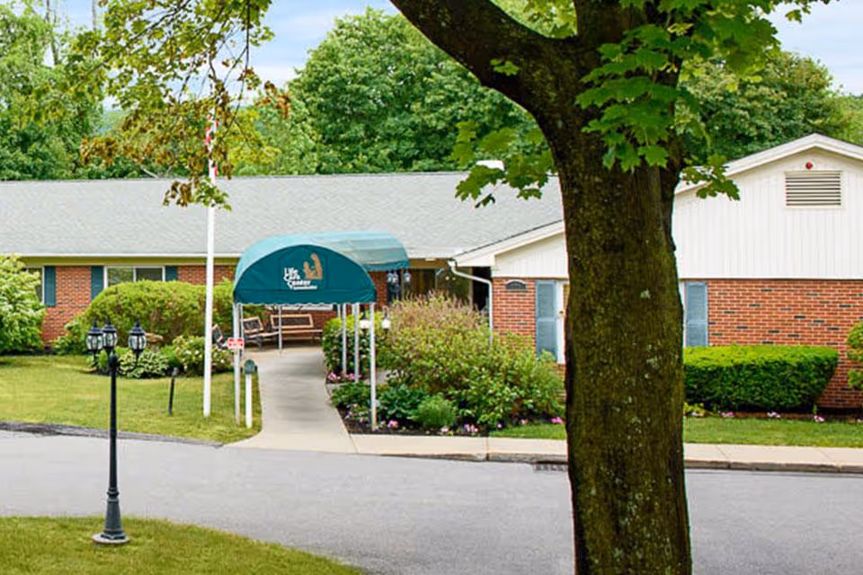 Front entrance of a brick senior living facility with a green awning, walkway, landscaping, and a large tree in the foreground.