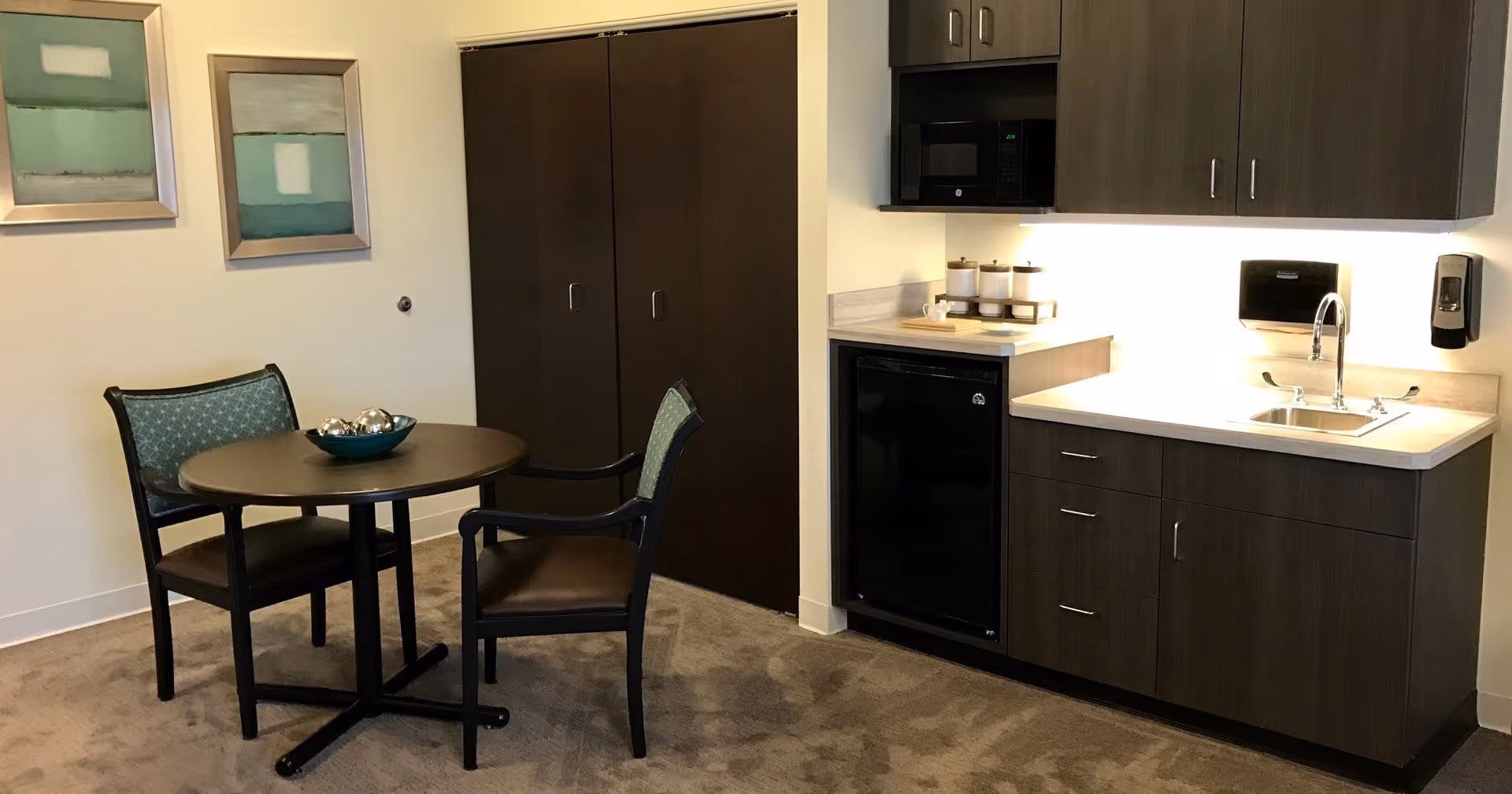 Small kitchenette with dark cabinets, countertop and sink beside a round dining table and two chairs, with wall art and a closed closet.