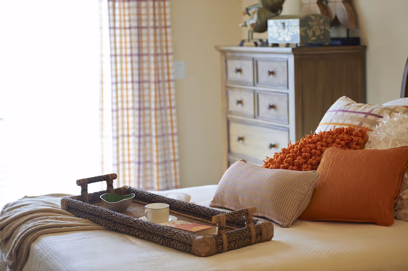 A cozy bedroom scene with a neatly made bed featuring multiple decorative pillows in shades of orange and beige. A woven tray with a cup and small bowl rests on the bed. In the background, there is a wooden dresser and a window with plaid curtains allowing natural light to fill the room.